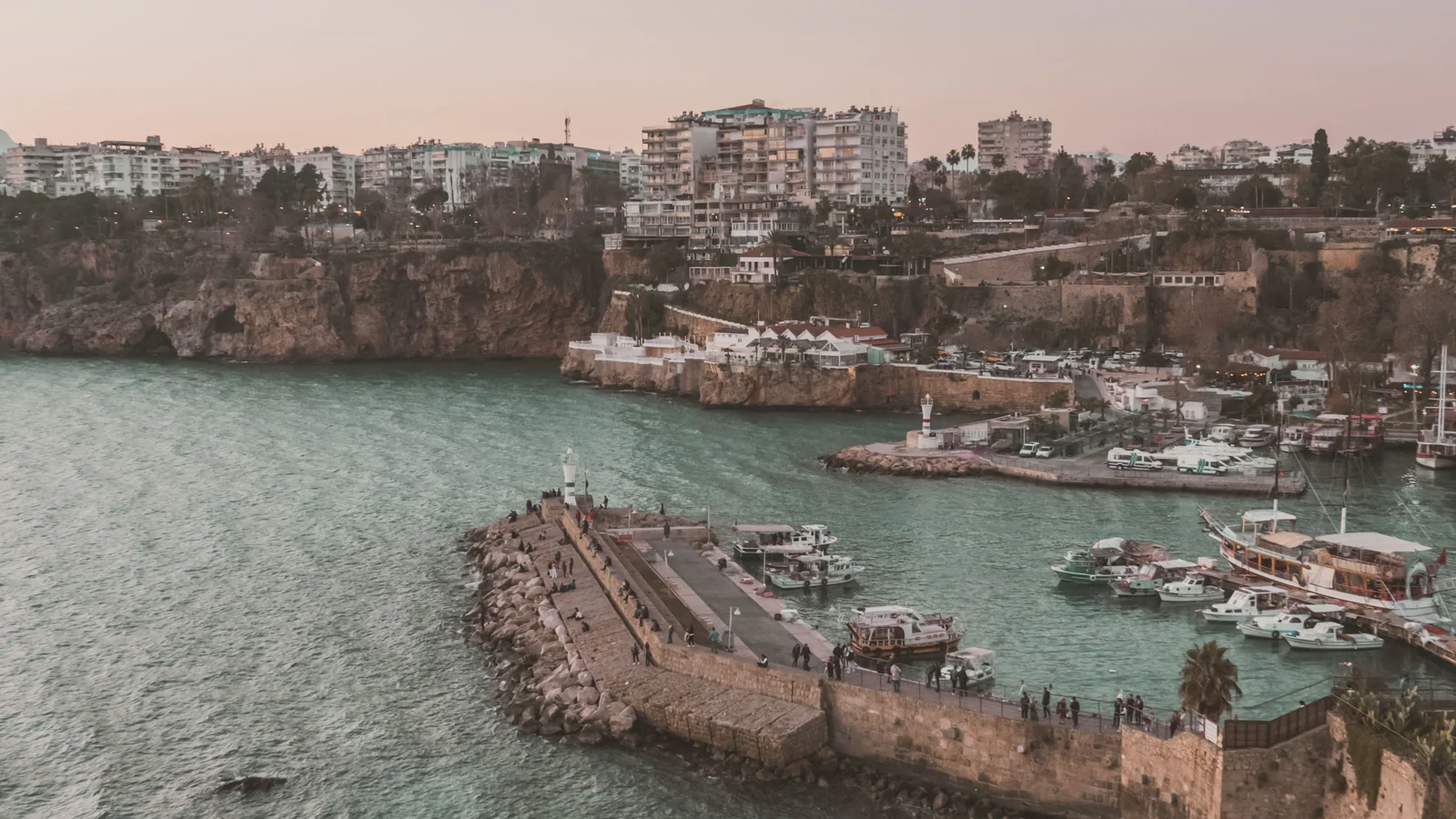 Antalya’s old harbour and stone breakwater beneath the city’s cliff-edge buildings at dusk.