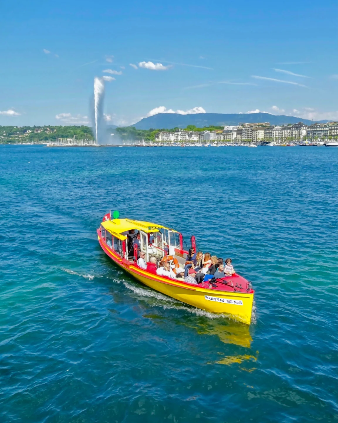 A yellow and red Mouettes shuttle boat on Geneva’s lake with the Jet d’Eau behind