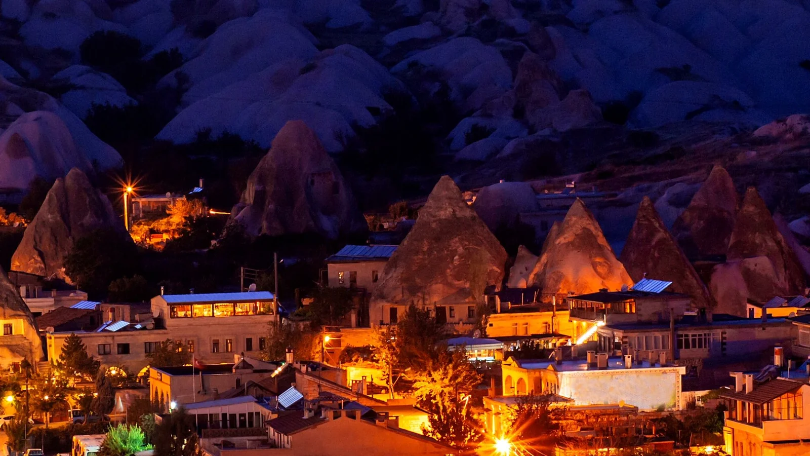 Illuminated stone houses, rock cones and a curving road on Göreme’s hillside after dusk.