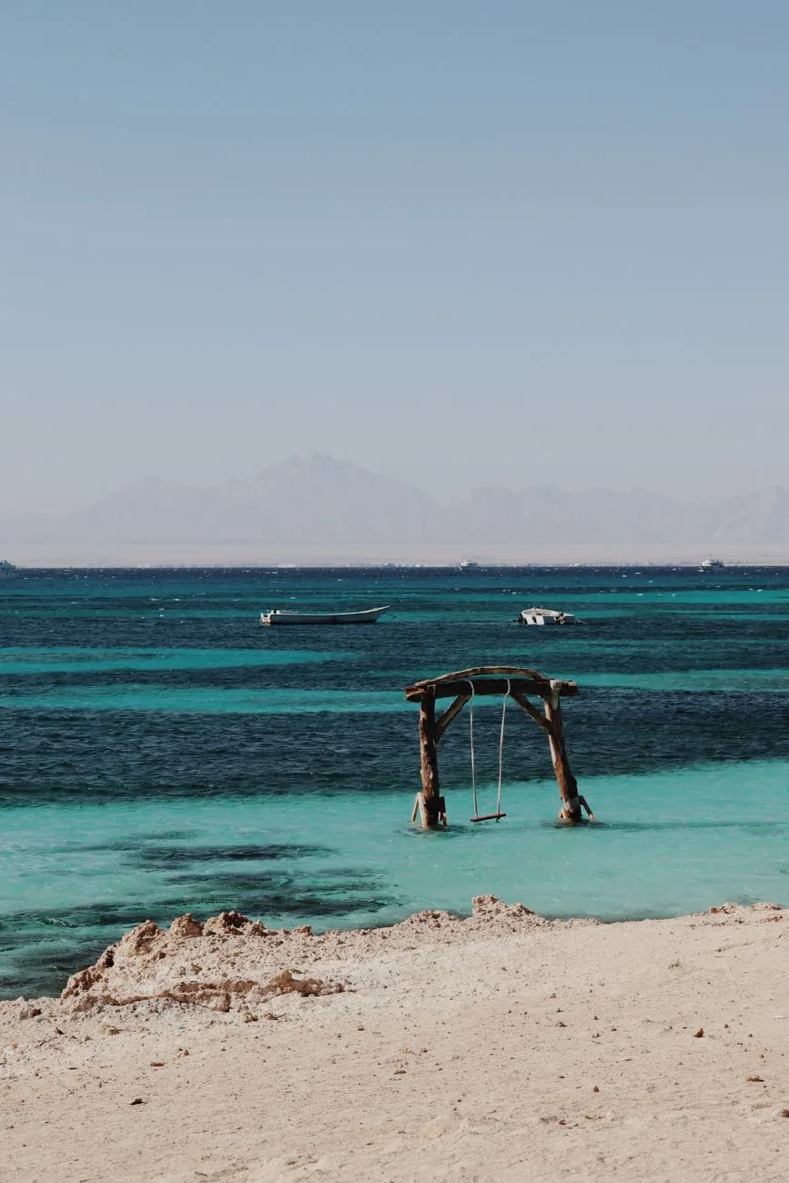 A wooden swing standing in shallow turquoise Red Sea water off Hurghada, with boats and distant mountains