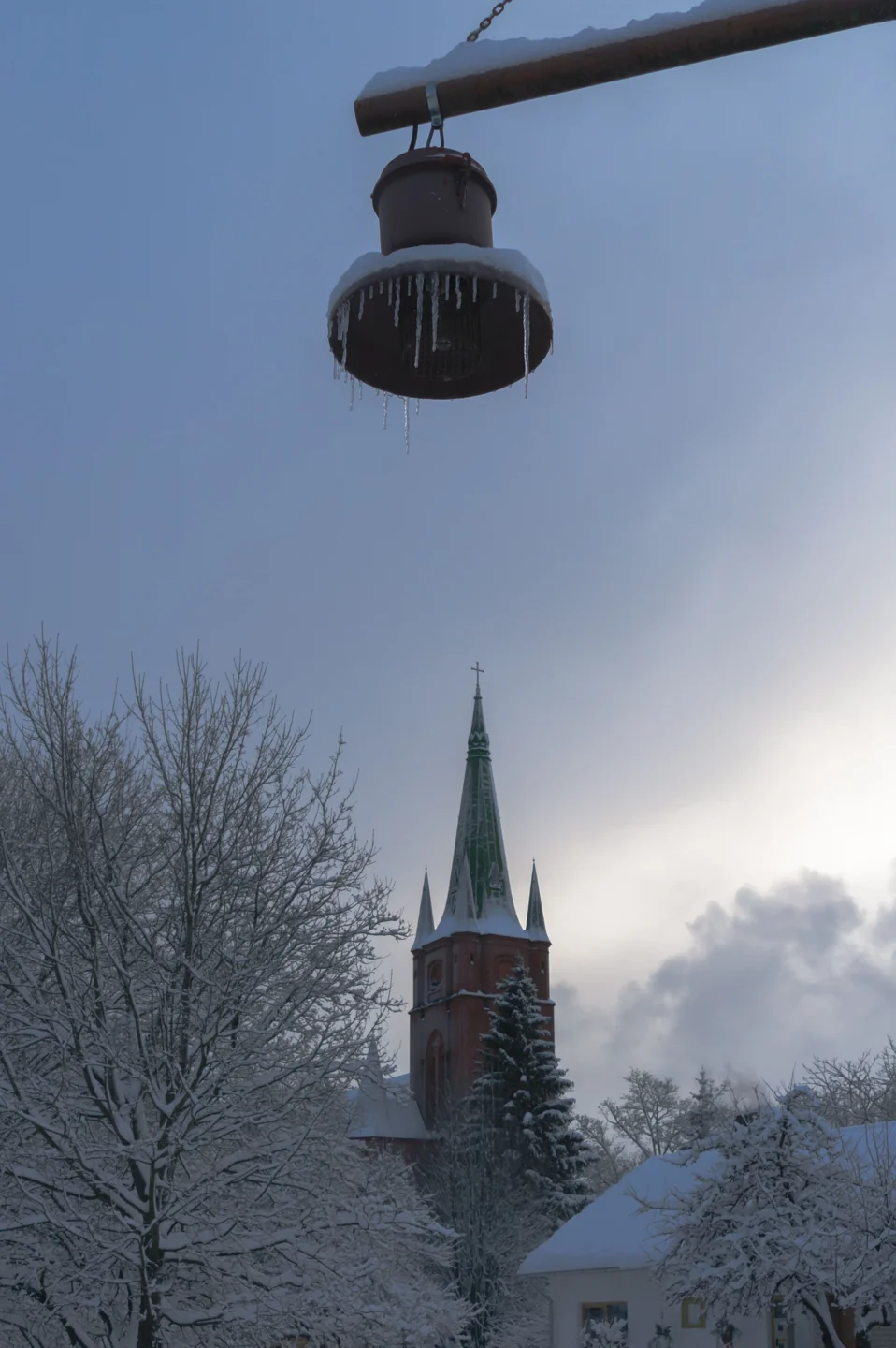 St Anne’s red-brick church tower and green spire behind snowy trees in Kuldīga, with an icy lamp in the foreground