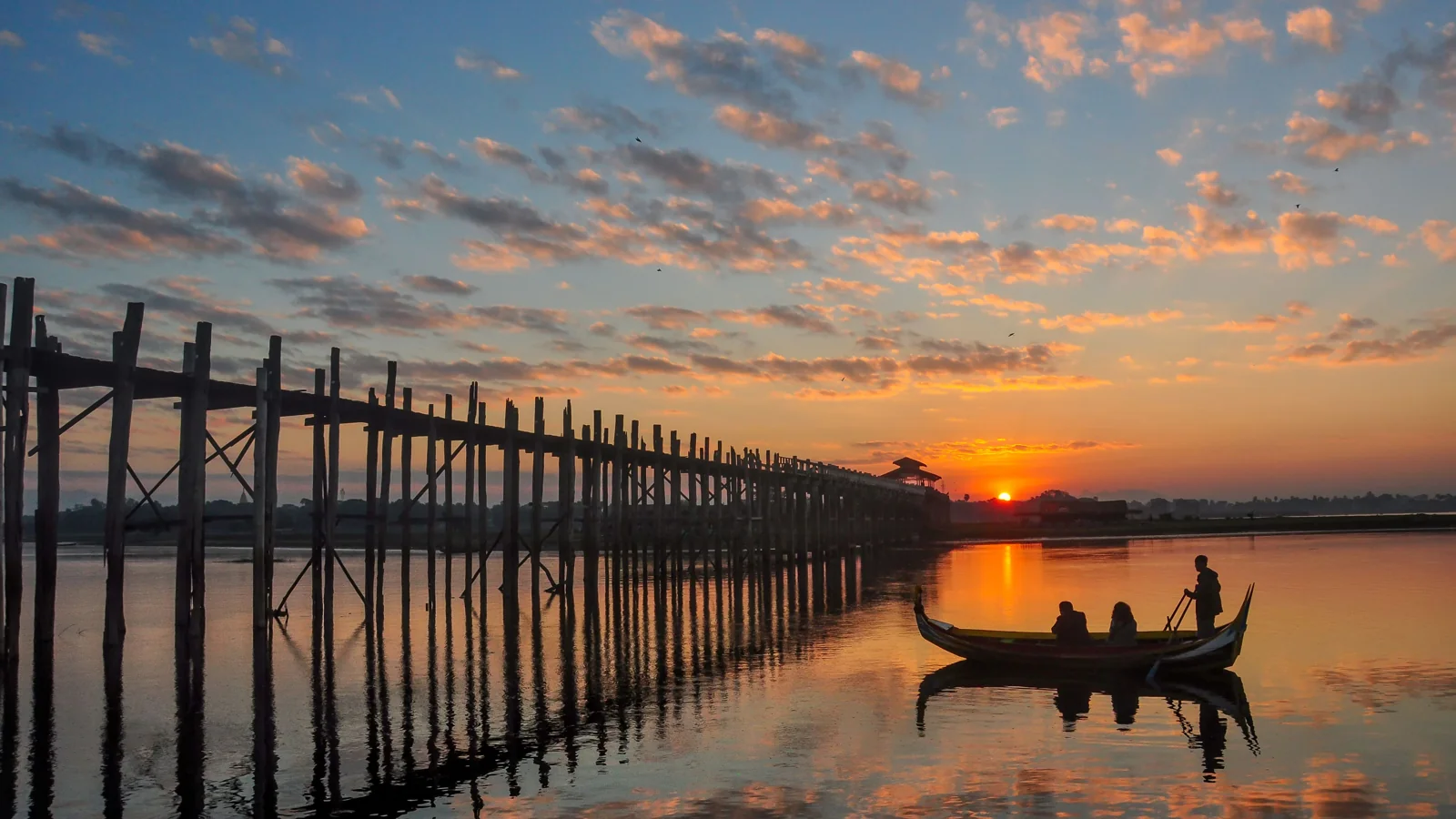 U Bein Bridge crossing Taungthaman Lake at Amarapura, with a small boat and low sun reflected on the water.
